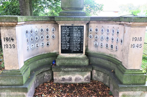 Ein steinernes Denkmal in einem eingezäunten jüdischen Friedhof in Berlin, Deutschland, mit eingravierten Text und Zahlen an seiner Wand, umgeben von Bäumen und verstreuten trockenen Blättern.