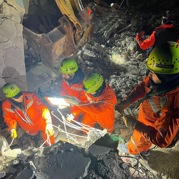 Eine Gruppe von Rettungskräften in orangefarbenen Uniformen und grünen Helmen räumt Trümmer von einem eingestürzten Gebäude weg, wobei sie Werkzeuge verwenden und ein Kran im Hintergrund zu sehen ist.
