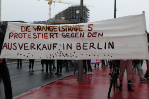 Eine Gruppe von Menschen steht auf der Straße und hält ein Transparent mit der Aufschrift "Die Wrangellstraße 5 protestiert gegen den Verkauf in Berlin", mit Fahrrädern, Pfählen, Gebäuden und einem Kran im Hintergrund unter einem sichtbaren Himmel.