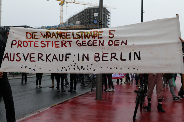 Eine Gruppe von Menschen steht auf der Straße und hält ein Transparent mit der Aufschrift "Die Wrangellstraße 5 protestiert gegen den Verkauf in Berlin", mit Fahrrädern, Pfählen, Gebäuden und einem Kran im Hintergrund unter einem sichtbaren Himmel.