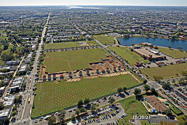 Luftaufnahme einer Stadt mit Gebäuden, Bäumen, Gras, Straßen mit Fahrzeugen, Wasser und bewölktem Himmel, mit einem großen zentralen Feld, das als Parkplatz vorgeschlagen ist.