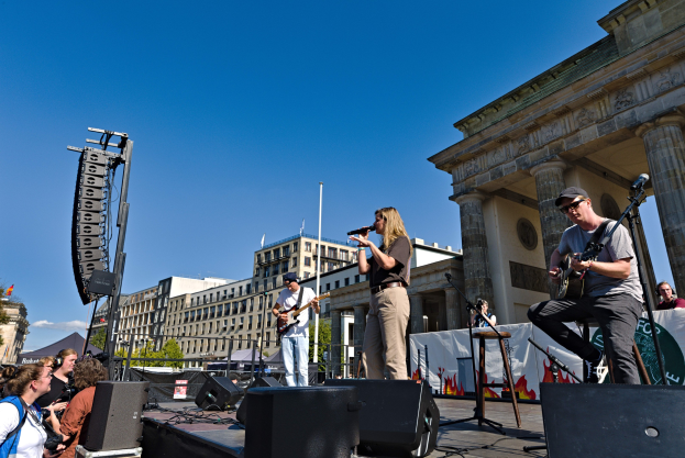 Eine Gruppe von Menschen, die auf einer Bühne vor dem Brandenburger Tor in Berlin Musik machen, mit Lautsprechern, Instrumenten und Mikrofonen gegen den Hintergrund von Gebäuden, Bäumen und einem klaren Himmel