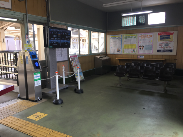 Ein belebter Bahnhofseingang mit Reihen von Stühlen, einer Service-Theke, Ticketautomaten, Kettenzaunpfählen, Informationsanzeigen, Wandplakaten, Deckenbeleuchtung und Geländern.