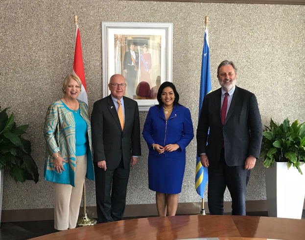 Gruppe von Menschen, die um einen Tisch mit Papieren stehen, umgeben von Fahnen, Topfpflanzen und einem gerahmten Foto an der Wand während einer Unterzeichnungszeremonie.