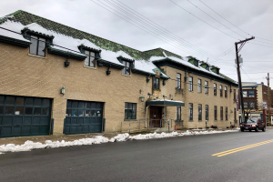 Großes Backsteingebäude mit grüner Dachfläche an einer Straßenkreuzung, umgeben von Fahrzeugen, Schnee, Strommasten, Bäumen und einer bewölkten Himmel, identifiziert als die ehemalige Feuerwache St. Louis.