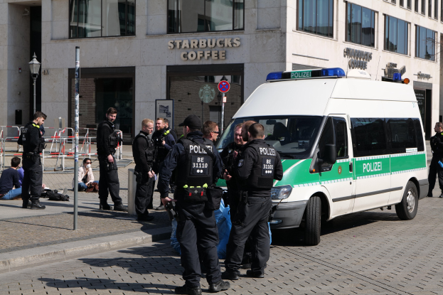 Polizeibeamte vor einem Starbucks-Café mit einem Van auf der rechten Seite und Menschen auf der linken Seite, vor einem Hintergrund aus einem Gebäude, einem Schild, einem Laternenmast und einem Zaun.