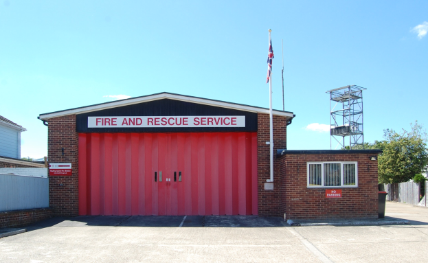 Feuerwehr- und Rettungsdienstgebäude mit roter Tür, Fenstern, einem Namenstafel, einem Fahnenmast mit einer Fahne, einem Metallturm, einem Zaun, einer Gruppe von Bäumen und einem bewölkten Himmel.