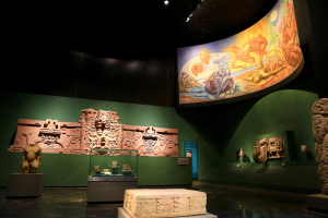 Ein Museumsraum mit verschiedenen Artefakten, darunter Skulpturen, Tischgegenstände, eine Texttafel, eine Wandmalerei und Deckenlampen im Anthropologischen Museum von Mexiko-Stadt.