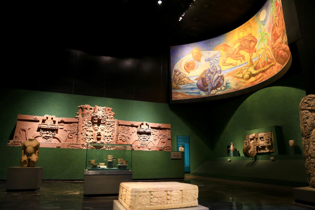 Ein Museumsraum mit verschiedenen Artefakten, darunter Skulpturen, Tischgegenstände, eine Texttafel, eine Wandmalerei und Deckenlampen im Anthropologischen Museum von Mexiko-Stadt.