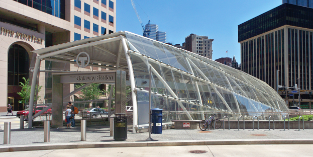 Eine belebte Stadtstraße mit hohen Wolkenkratzern, ein Bushaltestelle mit Glasdach, Menschen, Fahrzeuge, ein Metallzaun, ein Mülleimer, Pfosten, ein Schild, eine Ampel, Bäume, ein Kran und ein bewölkter Himmel.
