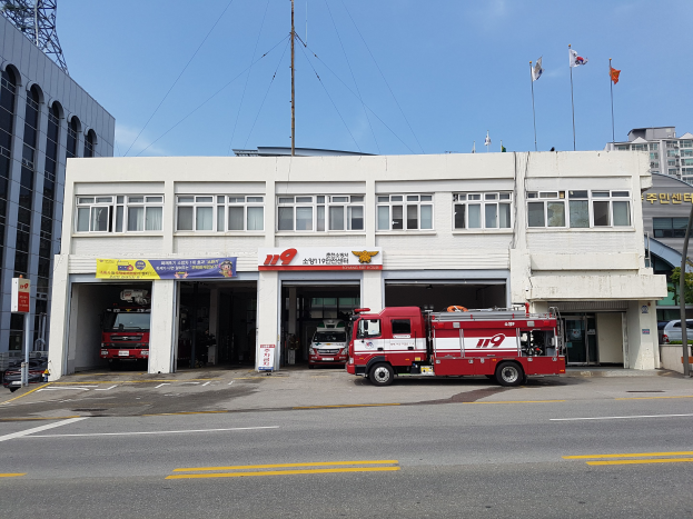 Feuerwache mit mehreren Feuerwehrautos davor geparkt, ein Gebäude mit Fenstern, Bannern und Fahnen unter einem bewölkten Himmel mit einem Turm links.