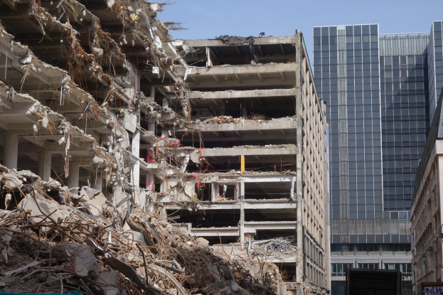 Ein große Abriss in einer Stadtumgebung, umgeben von intakten Gebäuden, Straßeninfrastruktur, Fahrzeugen, Bäumen und einem klaren Himmel.