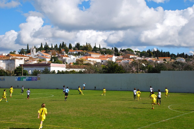 Gruppe von Menschen, die draußen ein Spiel spielen, mit Plakaten an der Wand, Bäumen, Strommasten, Häusern und Wolken im Hintergrund.