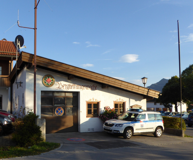 Polizeiauto vor einem Gebäude mit Fenstern und Türen geparkt, umgeben von Grünflächen einschließlich blühender Pflanzen, Gras und Bäumen, mit sichtbaren Straßenlaternen, einer Schüsselantenne und einem Schild unter einem bewölkten Himmel.