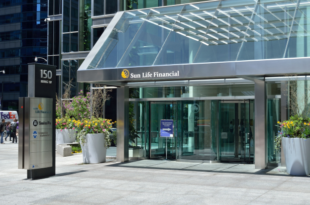 Moderner Glasbau der Sun Life Financial in Toronto mit einer Hinweistafel, Topfpflanzen mit Blüten, Menschen, einer Straßenlaterne, einer Hinweistafel, Fahrzeugen auf der Straße und einem bewölkten Himmel.