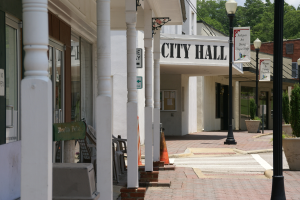 Ein Rathaus umgeben von Gebäuden, Straßenmasten, Straßenlaternen, Namensschildern, Kraftfahrzeugen, Verkehrskegeln, Stühlen, Topfpflanzen, Bäumen und einer bewölkten Himmel.