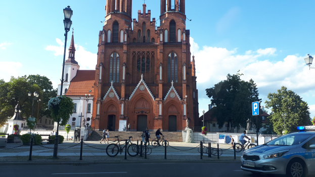 Polizeiauto vor einer Kirche mit umliegenden Gebäuden, Straßeninfrastruktur, Fahrzeugen, Radfahrern und Bäumen unter einem bewölkten Himmel.
