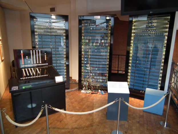 Ein unordentlicher Raum mit verschiedenen elektronischen Geräten, ein Tisch mit Gegenständen, eine wandhohe Tafel mit Text, Seilabsperrungen auf Stangen, ein Metallzaun, gerahmte Fotos an der Wand und Deckenleuchten.