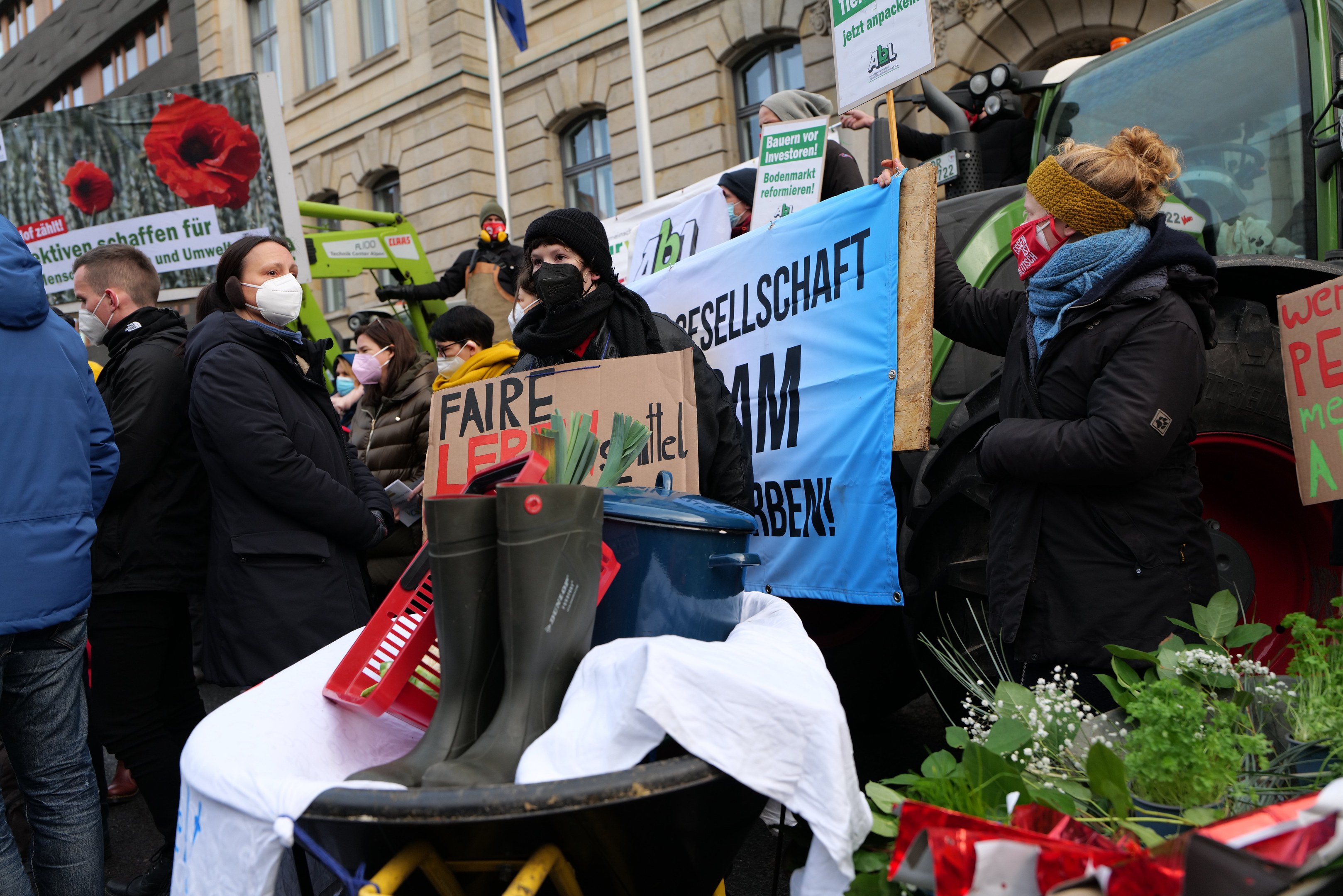 Eine Gruppe von Menschen mit Masken und Protestschildern vor einem Lastwagen, mit einem Tisch mit Gegenständen, Topfpflanzen und einem Flaggengebäude im Hintergrund.