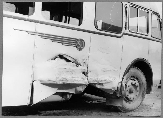 Ein Schwarz-Weiß-Foto eines beschädigten Busses am Straßenrand, mit eingedellter Front, zersplitterten Fenstern und einer offenen Fahrertür, die das Innere zeigt.