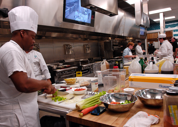 Eine Gruppe von Köchen in einer Küche, die Essen auf einem Schneidbrett zubereiten, mit einem Koch im Vordergrund in einer weißen Schürze und Mütze, der ein Messer hält und von Utensilien, Schüsseln, Gemüse, einem Handy, Flaschen und anderen Gegenständen umgeben ist, während im Hintergrund ein Fernseher und Deckenlampen zu sehen sind.