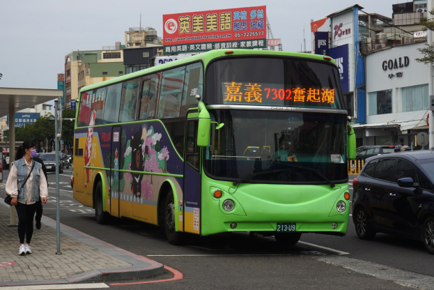 Ein grüner Bus fährt auf einer Straße mit hohen Gebäuden, eine Frau geht auf dem Gehweg und trägt eine Tasche, Bäume und Pfosten säumen die Straße, und ein klarer blauer Himmel ist zu sehen.