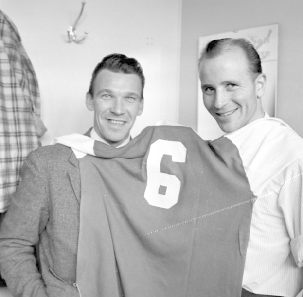 Zwei Männer in Schwarz und Weiß stehen nebeneinander und lächeln, während sie ein Fußballtrikot hochhalten; im Hintergrund ist eine Wand mit einer Tafel und einem Haken zu sehen.