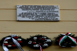 Zwei Kränze mit Bändern hängen an der Seite eines Gebäudes, dahinter eine Gedenktafel für Holocaust-Opfer.