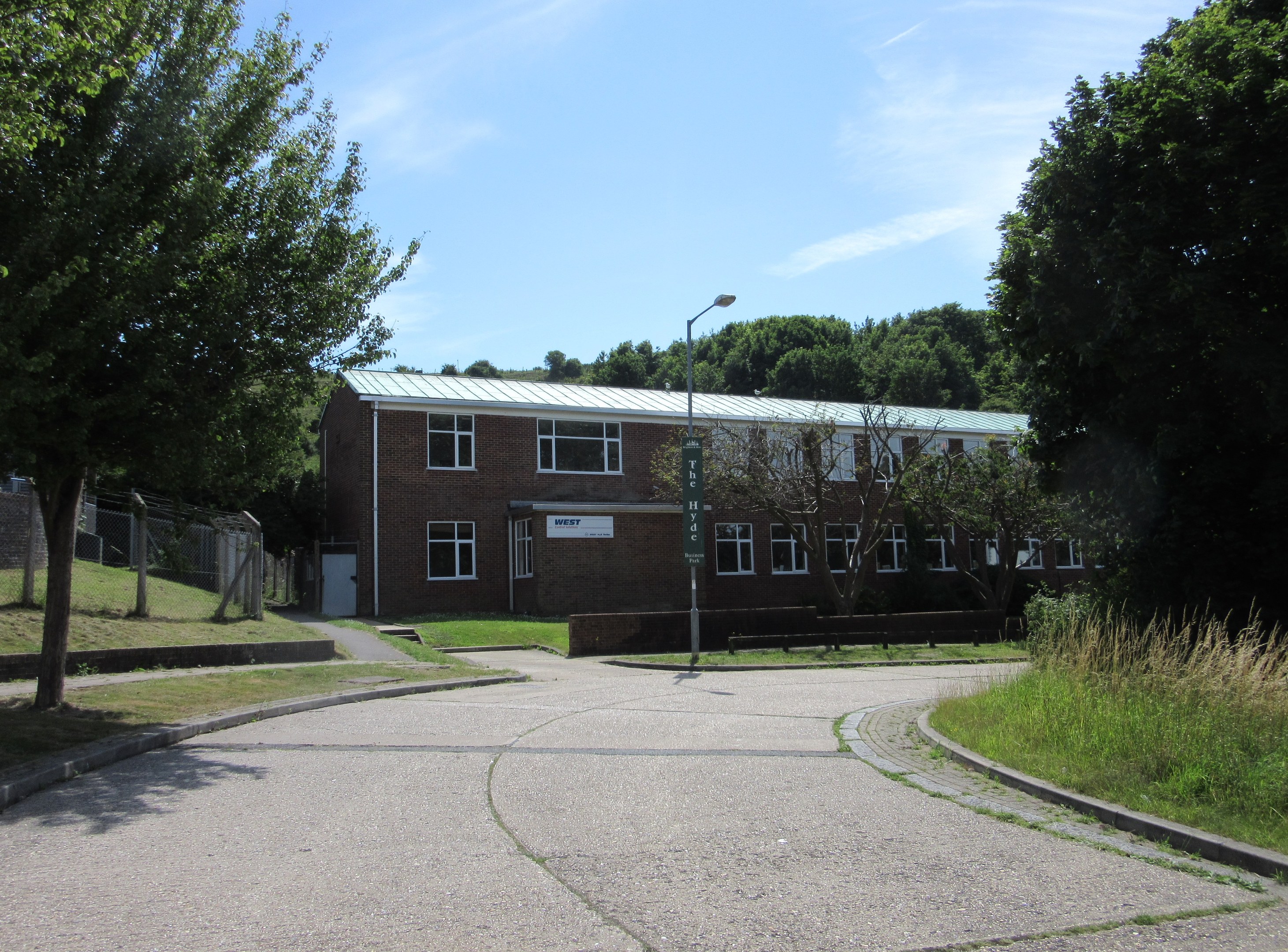 Ein Schulgebäude mit Fenstern, einem Straßenpfahl, einem Schild, einer Gruppe von Bäumen, Gras, einem Zaun und einem bewölkten Himmel, das in der Mitte eines Parks umgeben von saftigem Gras liegt.