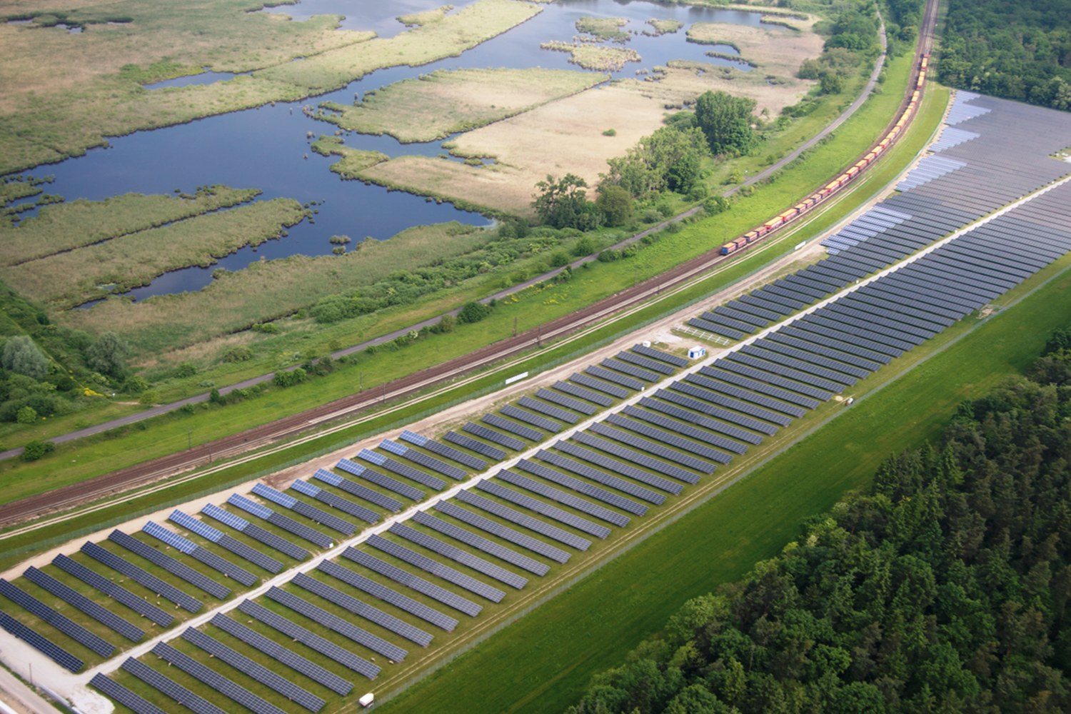 Luftaufnahme einer Solar-Farm mit Panelen, umgeben von Bäumen, Gras, Wasser und einer nahen Bahntrasse.