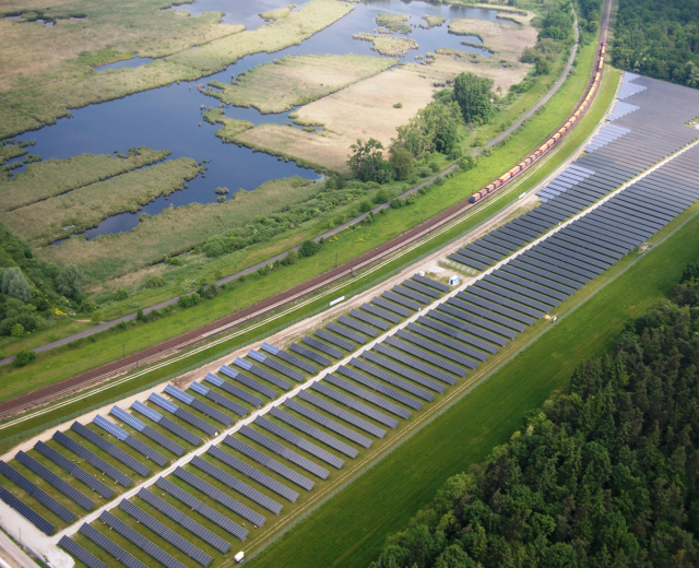 Luftaufnahme einer Solar-Farm mit Panelen, umgeben von Bäumen, Gras, Wasser und einer nahen Bahntrasse.