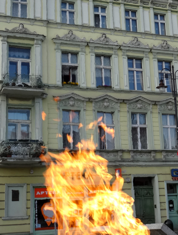 Ein großes Feuer brennt vor einem Gebäude mit Fenstern und Türen, einem Laternenmast und einer Tafel mit Schrift darauf.