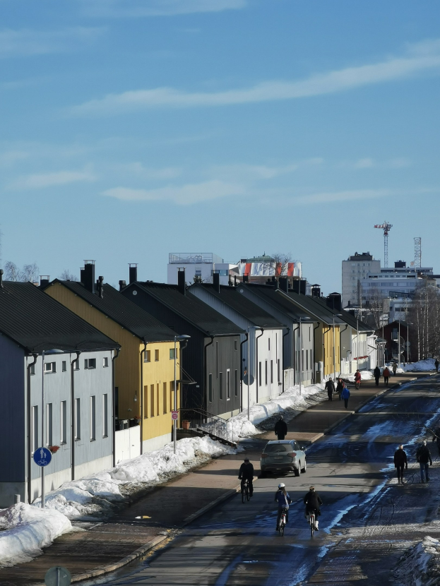 Eine Gruppe von Menschen, die auf Fahrrädern eine schneebedeckte Straße entlangfährt, gesäumt von Gebäuden, Bäumen, Pfählen und Schildern unter einem bewölkten Himmel.