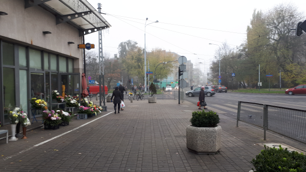 Eine Stadtstraße mit Menschen auf dem Gehweg, Fahrzeugen auf der Straße und verschiedenen städtischen Elementen wie Bäumen, Pfosten, Laternen und Bänken.