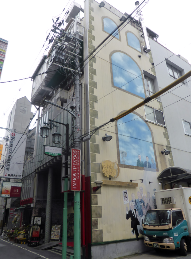 Ein Lastwagen steht auf der Straßenseite neben einem hohen Gebäude mit einer Wandmalerei, mit Strommasten, Kabeln, Straßenlaternen, Namensschildern, Kraftfahrzeugen, Topfpflanzen und einem klaren blauen Himmel im Hintergrund.