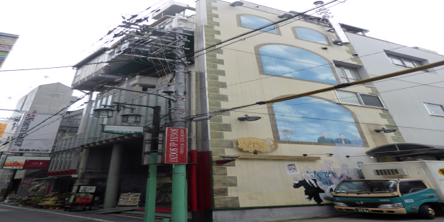 Ein Lastwagen steht auf der Straßenseite neben einem hohen Gebäude mit einer Wandmalerei, mit Strommasten, Kabeln, Straßenlaternen, Namensschildern, Kraftfahrzeugen, Topfpflanzen und einem klaren blauen Himmel im Hintergrund.