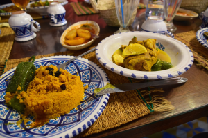 Ein Tisch mit Tellern voller Essen, Gläsern mit Wein, Schalen, Gefäßen, Besteck und Matten, mit Menschen im Hintergrund bei einem traditionellen marokkanischen Mahl.