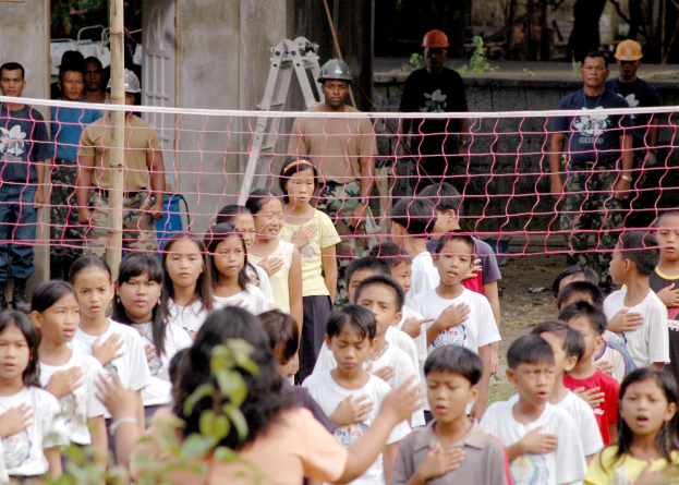 Gruppe von Kindern vor einem Netz mit einer Frau im Vordergrund; Hintergrund enthält Personen mit Helmen, eine Leiter, eine Wand und Bäume.