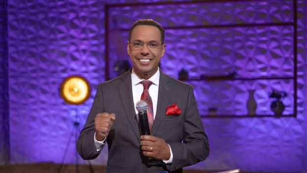 Stephen Colbert, Moderator von "The Daily Show with Trevor Noah", steht auf einer Bühne und hält ein Mikrofon in der Hand, trägt einen Anzug, eine Krawatte und eine Brille, hinter ihm ist ein Scheinwerfer und ein Ständer zu sehen.