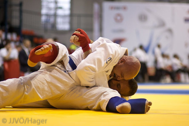 Zwei Judoka im Kampf, mit Zuschauern und Innenraum-Elementen im Hintergrund.