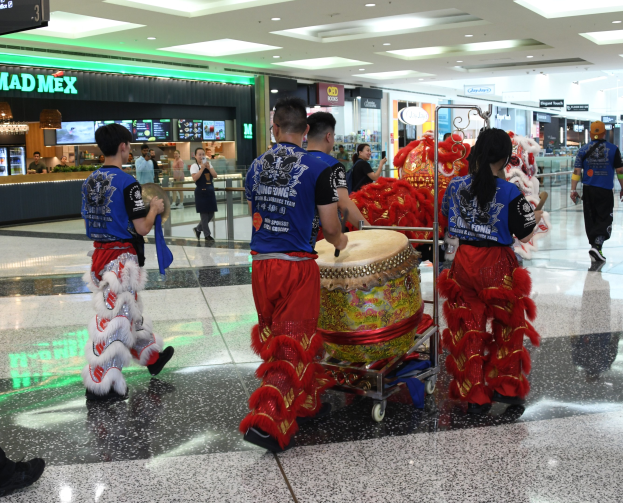 Eine Gruppe von Menschen in roter und blauer Kleidung mit Musikinstrumenten und Wagen geht durch einen hell erleuchteten Flughafen während einer Chinesischen Neujahrsfeier in Singapur.