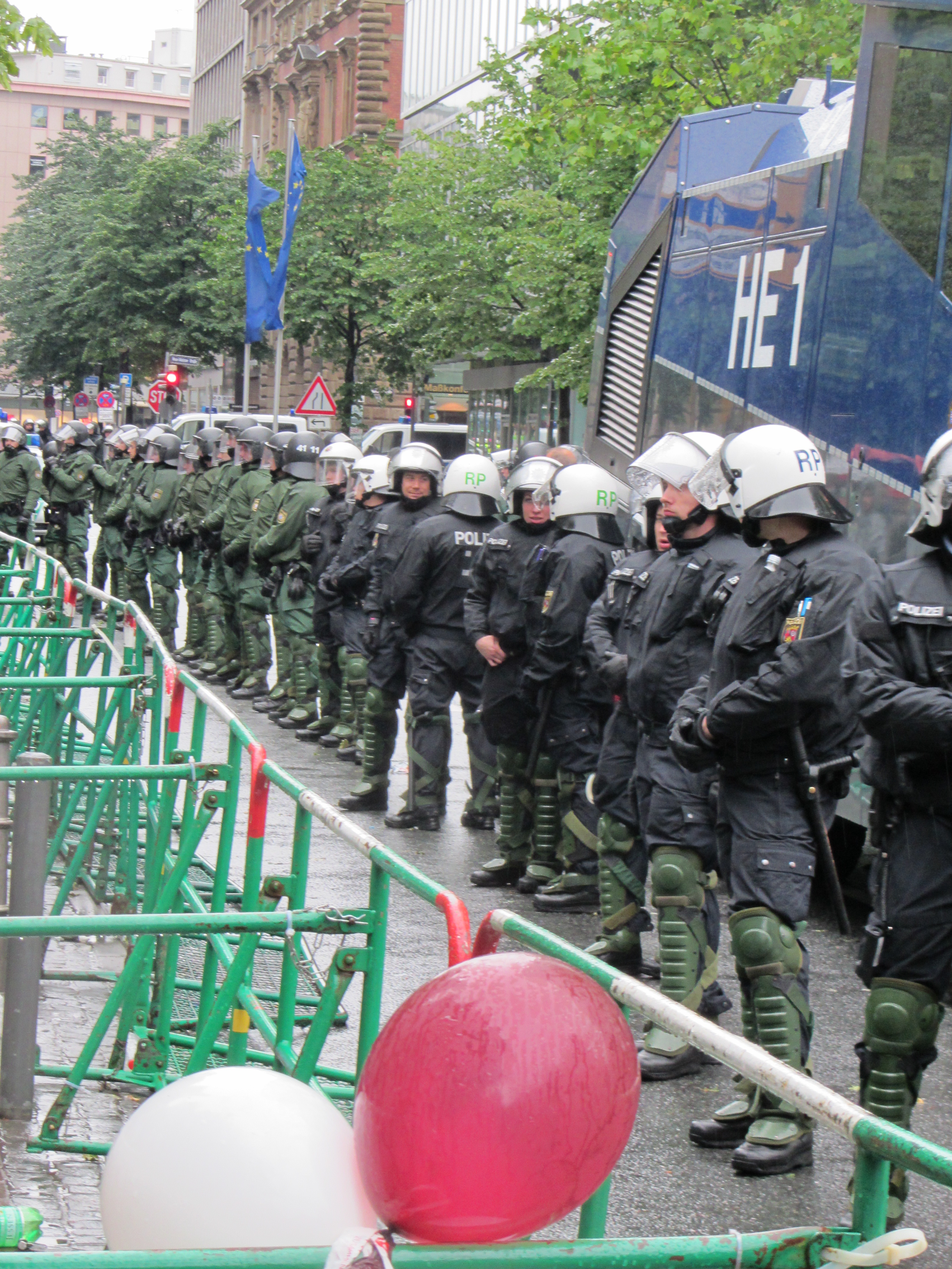 Eine Gruppe von Polizisten in Helmen und mit Waffen in der Hand steht vor einer Absperrung auf einer Straße mit Geländern und Luftballons, mit Bäumen, Gebäuden mit Fenstern, Fahnen und einem klaren blauen Himmel im Hintergrund.