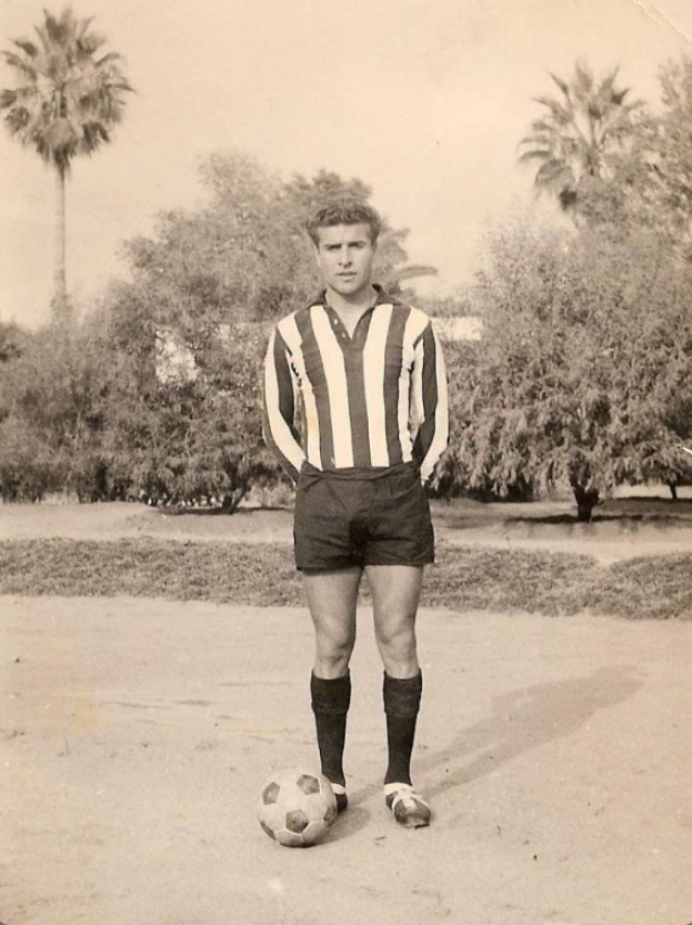 Schwarzes und weißes Foto eines Mannes, der auf Gras steht und einen Fußball hält, mit Bäumen und Himmel im Hintergrund.
