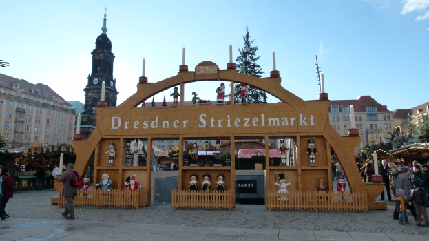 Ein geschäftiger Weihnachtsmarkt in Dresden, Deutschland, mit Menschen, die gehen, einige mit Taschen, unter einem Torbogen, mit Bäumen, Gebäuden und bewölktem Himmel im Hintergrund.
