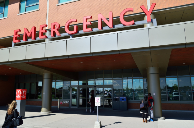 Eine Gruppe von Menschen, die vor einem Notfallgebäude mit Glaswänden und Säulen, einem Namensschild, Hinweistafeln auf Pfählen und einem Mülleimer gehen.