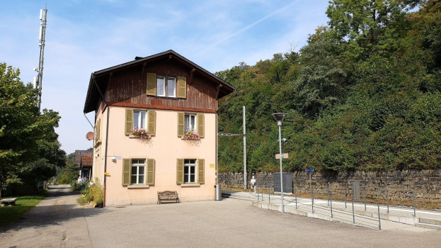 Ein mehrstöckiges Gebäude mit zahlreichen Fenstern, einem Vorgartenzaun und umgebenden Straßen-Elementen wie Pfählen, Laternen, Gittern, einer Bank, blühenden Pflanzen, Gras, Bäumen und einer Wand, unter einem bewölkten Himmel, gelegen in Baden-Württemberg, Deutschland.