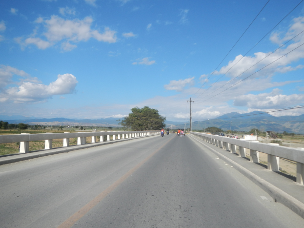 Autobahn mit Motorrädern, Geländern, Strommasten, Bäumen, Hügeln und einem bewölkten Himmel im Hintergrund.