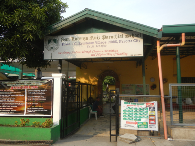 Der Eingang der San Lorenzo Ruiz Pfarrschule hat eine Tafel mit Text, einen Metallzaun, Menschen auf Stühlen unter einem Dach, Pfosten, ein Schild, Pflanzen, einen Baum und einen bewölkten Himmel.