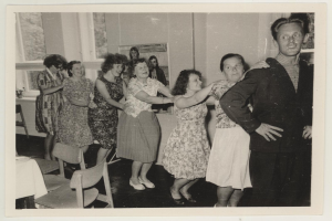 Eine Gruppe von Menschen tanzt in einem rückblickend 1950er-stiligen Raum mit Stühlen, einem mit einer wei├čen Decke bedeckten Tisch, einem Photo auf der Wand und Fenstern im Hintergrund.