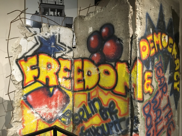 Wand mit Graffiti, das "Freiheit und Demokratie" in großen schwarzen Buchstaben liest, einem Geländer unten und einem Gebäude mit Fenstern im Hintergrund.